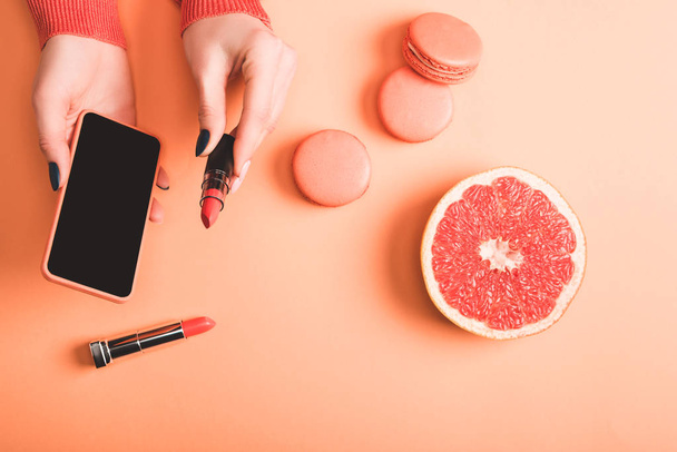 cropped view of woman holding smartphone and coral lipstick, macarons and grapefruit half on coral background, color of 2019 concept - Photo, Image