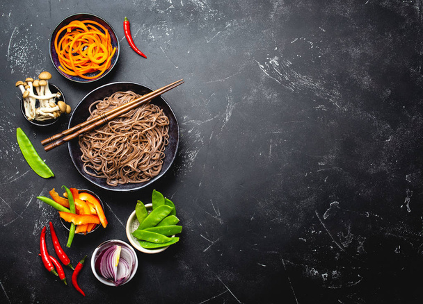 Ingredients for making stir-fried noodles soba. Cut fresh vegetables, mushrooms, boiled soba noodles in bowl with chopsticks ready for cooking, black stone background, space for text, top view - Фото, зображення
