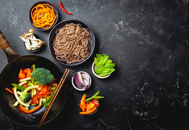Ingredients for making stir-fried noodles soba. Cut fresh vegetables in wok pan, boiled soba noodles in bowl with chopsticks ready for cooking, black stone background, space for text, top view - Photo, Image