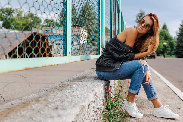 Fashion portrait of trendy young woman wearing sunglasses, jeans with halls and bomber jacket in the city. Copy space. - Foto, Imagem