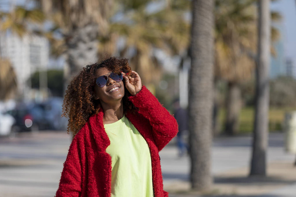 Front view of young beautiful curly woman standing on path smiling while looking camera in sunny day - Foto, afbeelding