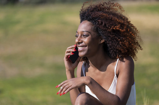 Front view of a young beautiful curly smiling woman sitting on ground in a city park while using a mobile phone outdoors. - Photo, Image