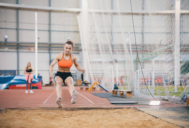 Молодая женщина с блондинистыми волосами прыгает через препятствие и улыбается в спортзале
 - Фото, изображение