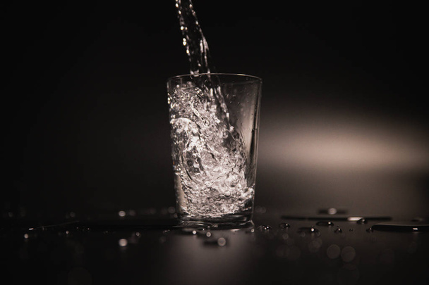 Water is poured into a transparent glass on a dark background - Foto, Bild
