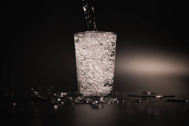 Water is poured into a transparent glass on a dark background - Zdjęcie, obraz