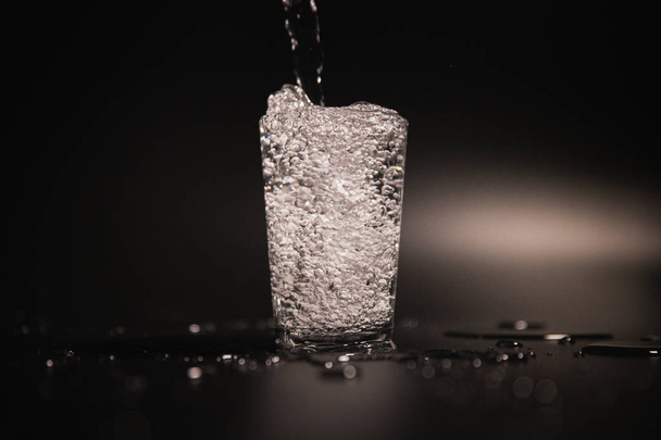 Water is poured into a transparent glass on a dark background - Φωτογραφία, εικόνα