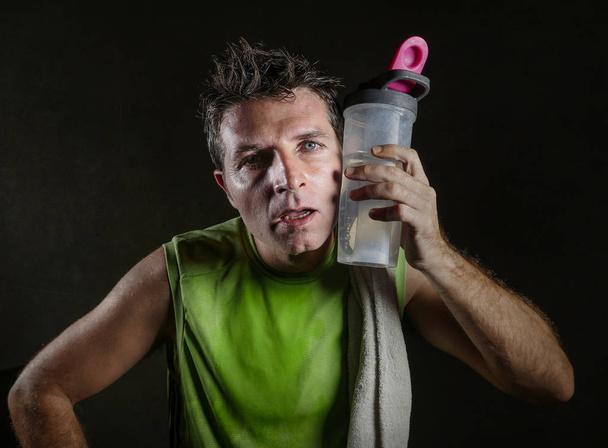 young tired and exhausted sport man with towel on his shoulder sweaty drinking water bottle after hard workout and fitness training isolated on dark background in healthy lifestyle concept - Foto, immagini