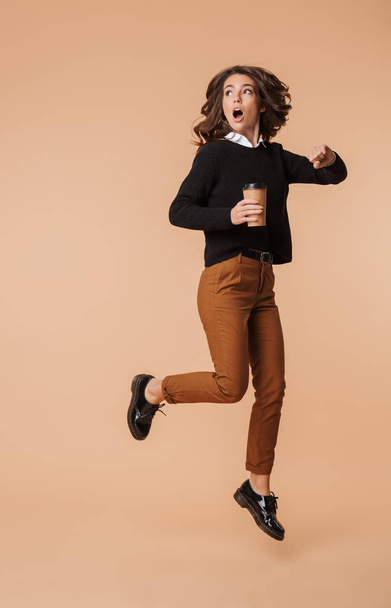 Shocked young woman wearing sweater running late isolated over beige background, holding takeaway coffee cup - Фото, зображення