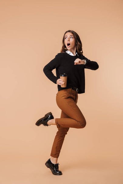 Shocked young woman wearing sweater running late isolated over beige background, holding takeaway coffee cup - Foto, Imagem