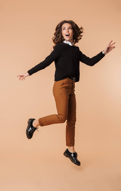 Full length of a smiling young woman wearing sweater jumping isolated over beige background - Fotó, kép