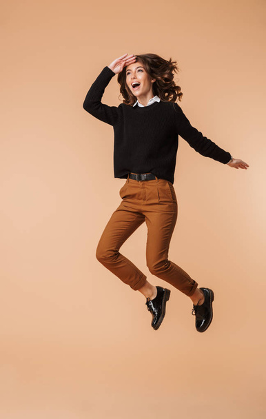 Full length of a smiling young woman wearing sweater jumping isolated over beige background - Foto, Imagen
