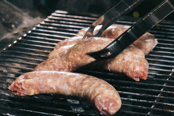 Close up van pincet in de buurt van ongekookte verse worst op BBQ-grill roosters - Foto, afbeelding
