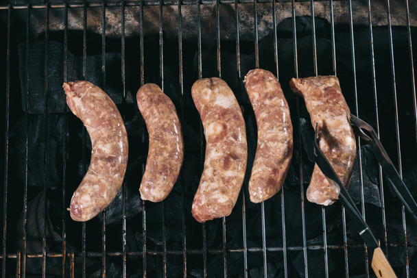 vue du dessus des pinces à épiler près des saucisses fraîches non cuites sur les grilles de barbecue
 - Photo, image