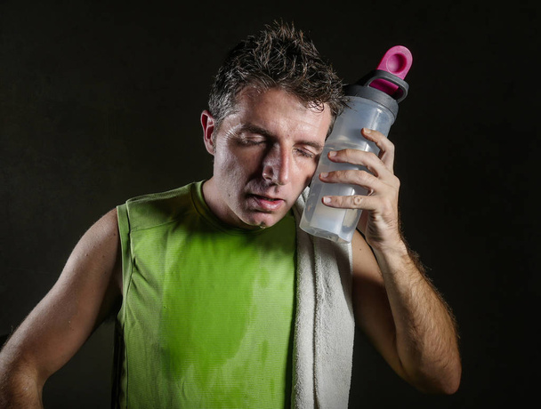 young tired and exhausted sport man with towel on his shoulder sweaty drinking water bottle after hard workout and fitness training isolated on dark background in healthy lifestyle concept - Foto, immagini