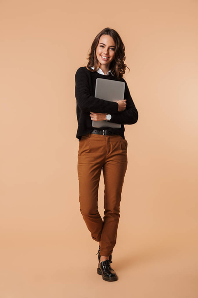 Photo of smiling woman 20s standing and holding laptop isolated over beige background - Valokuva, kuva