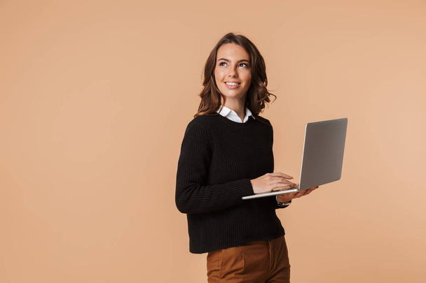 Photo of cute woman 20s standing and holding laptop isolated over beige background - Foto, imagen