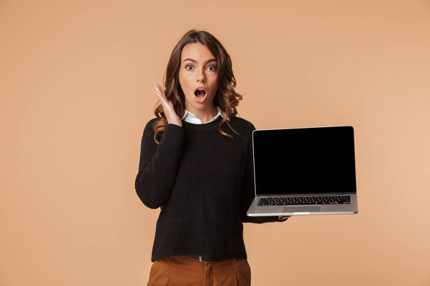 Happy young woman wearing sweater showing blank screen laptop computer isolated over beige background - Фото, зображення