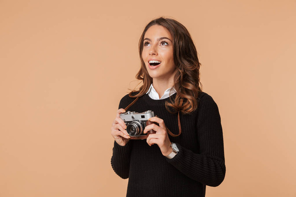 Pretty young woman wearing sweater holding photo camera isolated over beige background, looking away - Foto, immagini