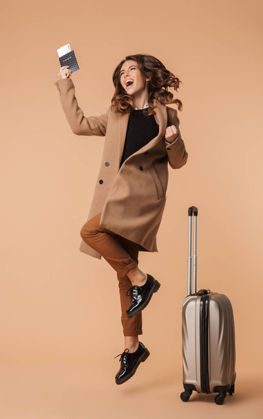 Full length of a cheerful young woman wearing a coat jumping with a travel bag isolated over beige background, showing passport with flight tickets - 写真・画像