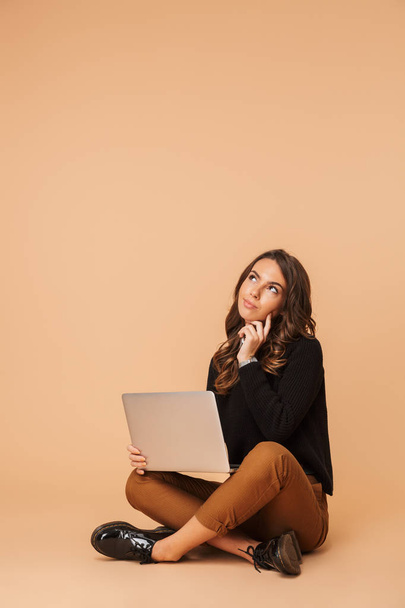 Image of beautiful woman 20s using laptop while sitting on floor isolated over beige background - Fotoğraf, Görsel