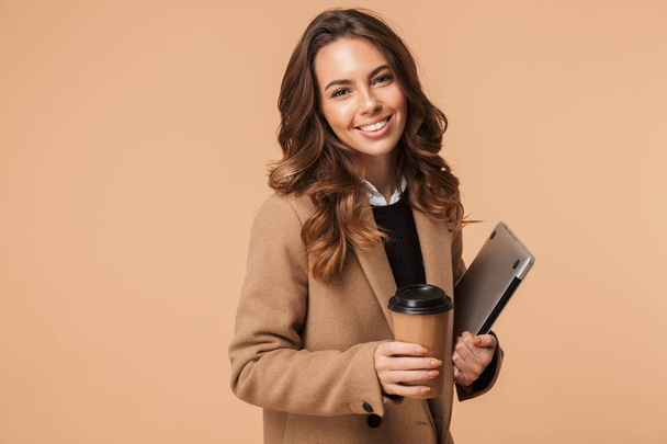 Smiling young woman wearing a coat standing isolated over beige background, carrying laptop and drinking coffee - Zdjęcie, obraz
