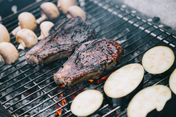 selective focus of juicy tasty steaks grilling on hot coals with mushrooms and sliced eggplant - Fotó, kép