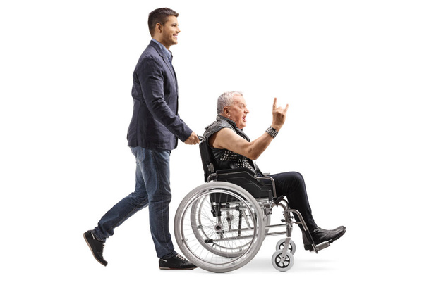 Full length profile shot of a young man pushing an man in leather vest making a rock and roll hand sign in a wheelchair isolated on white background - Photo, image