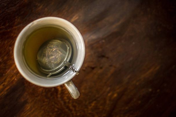 Чашка чая с infuser в горячей дымчатой воде с зеленым чаем, мятой травы и медом на темном деревянном фоне
 - Фото, изображение