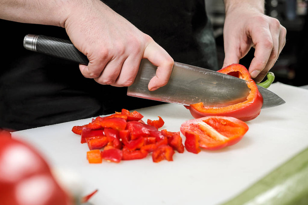 Male chef cuts vegetables for salad in a restaurant in a black apron. White cutting board, closeup of hands. - 写真・画像