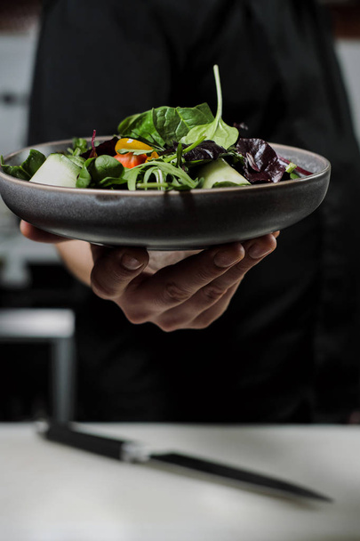Cook man in a black apron holds a dish of salad in his hand. Against the background of the kitchen of the restaurant. - Photo, image