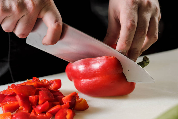 Male chef cuts vegetables for salad in a restaurant in a black apron. White cutting board, closeup of hands. - Φωτογραφία, εικόνα