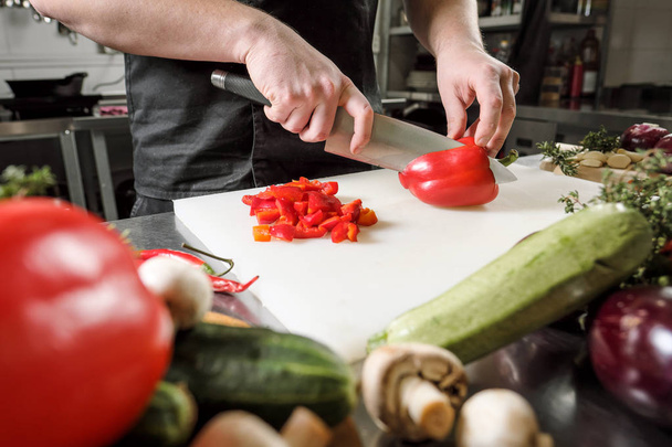 Male chef cuts vegetables for salad in a restaurant in a black apron. White cutting board, closeup of hands. - Foto, afbeelding