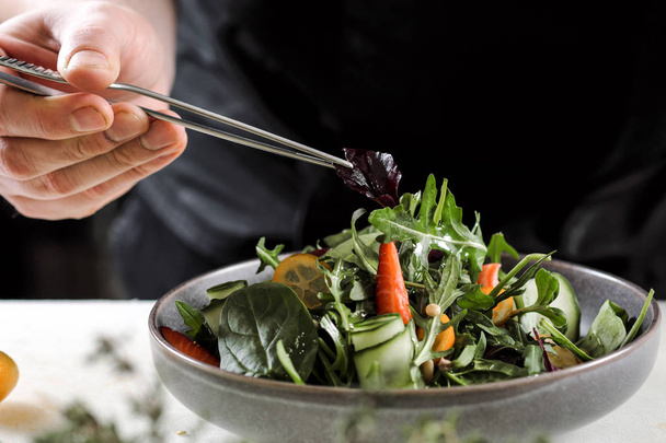 A male chef decorates a salad dish in a restaurant kitchen in a black apron. White cutting board. - Φωτογραφία, εικόνα