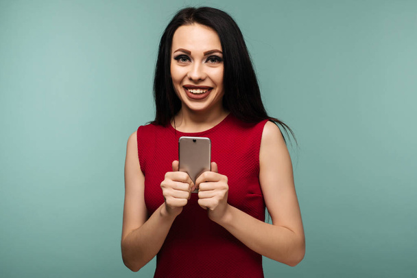Podekscytowany śmiejąca się kobieta w czerwonej sukience stoi i przy użyciu telefonu komórkowego na niebieskim tle - obraz - Zdjęcie, obraz