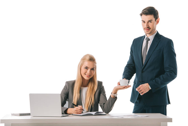 Красивая секретарша принесла чашку кофе для улыбающейся бизнесвумен на рабочем месте с ноутбуком, изолированным на белом
 - Фото, изображение