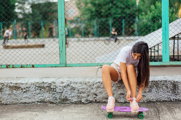 Summer lifestyle image of trendy pretty young girl sitting next to the skateboard coart with her purple plastic skateboard and tying shoelaces. - Foto, afbeelding