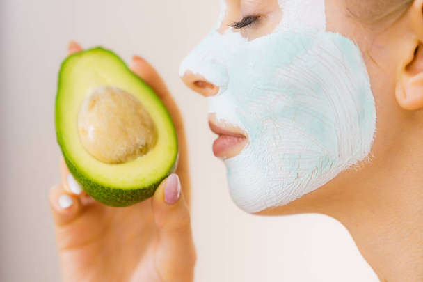 Young woman with white green mud mask on face holding avocado fruit. Teen girl taking care of her skin, cleaning the pores. Beauty treatment. - Фото, зображення