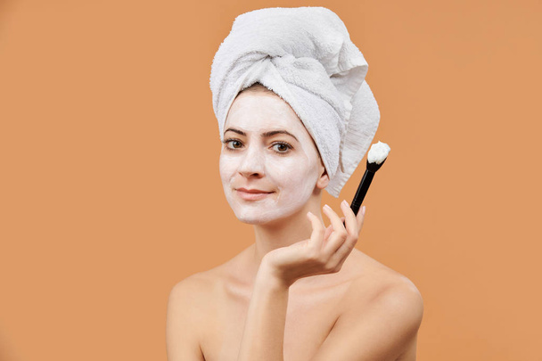 Young woman with white bath towel in her hair holding brush, applying face mask. Wellness and Spa concept on beige background. - Fotoğraf, Görsel