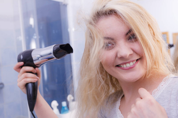Haircare. Beautiful long haired blonde woman drying hair in bathroom. Smiling girl blowing wind on wet head using holding hairdryer in hand. - Zdjęcie, obraz