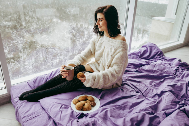 Młoda brunetka kobieta picia herbaty, kawy i jeść ciasteczka siedząc w pobliżu okna  - Zdjęcie, obraz