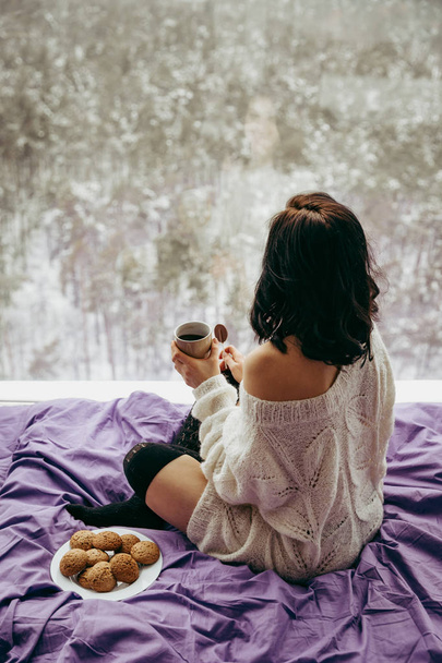 Młoda brunetka kobieta picia herbaty, kawy i jeść ciasteczka siedząc w pobliżu okna  - Zdjęcie, obraz