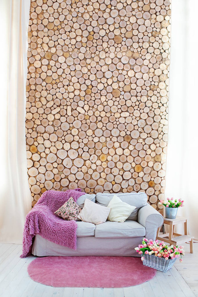ein helles Sofa mit Kissen und rosa Bezug, gegen die Wand aus gesägtem Holz. ein pinkfarbener Teppich am Boden. Blumen in einem Korb und einem Eimer. - Foto, Bild