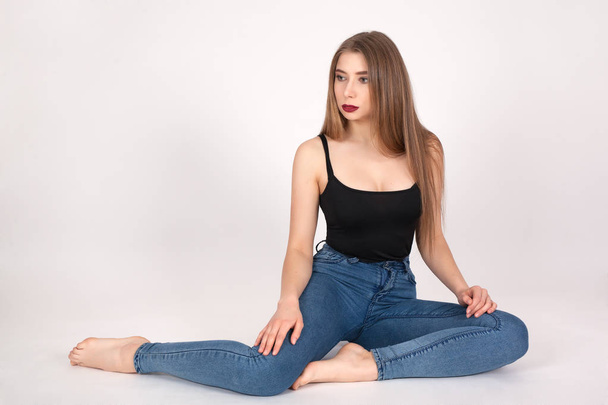 Portrait of a young beautiful girl with make-up isolated on white background. A woman in jeans and a t-shirt is sitting and looking sideways. - Photo, Image