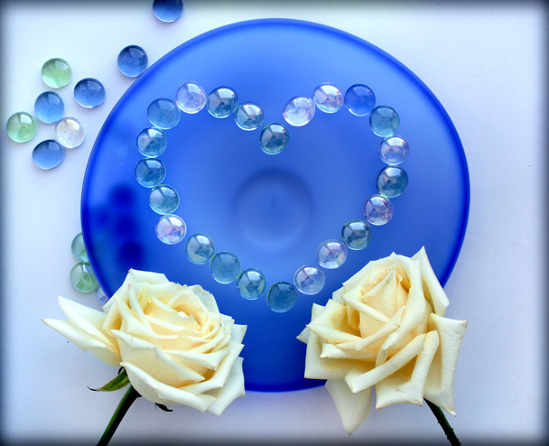 δύο άσπρα τριαντάφυλλα με μια καρδιά από γυάλινες χάντρες σε μπλε γυάλινο πιάτο - Φωτογραφία, εικόνα
