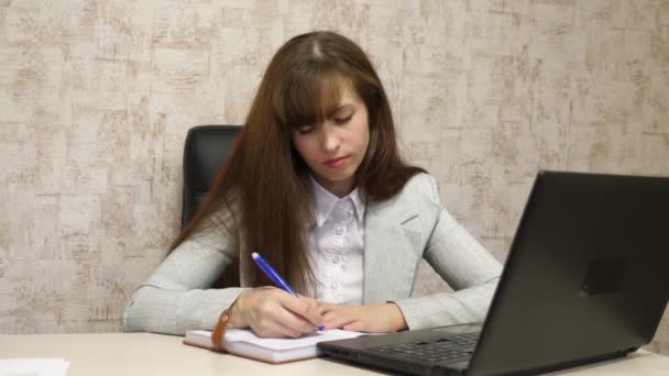 дівчина сидить у кріслі за комп'ютером і робить нотатки в блокноті. молода жінка-підприємець, що працює за ноутбуком. секретар пише ручку в блокноті
 - Кадри, відео