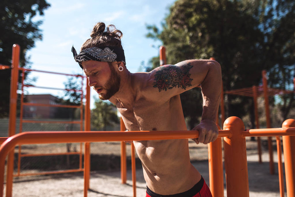 Young athletic man in headband  with a naked torso with tattoos doing push-ups on the uneven bars outside on a sunny day - Фото, изображение