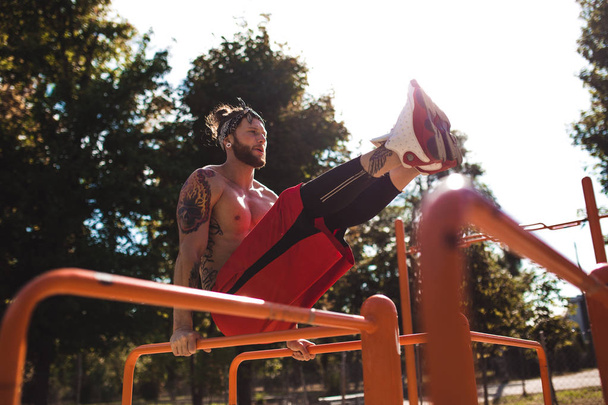 Young man in headband with naked torso dressed in black leggings and red shorts doing press exercises on the horizontal bar on the sports ground outside on a sunny day - Zdjęcie, obraz