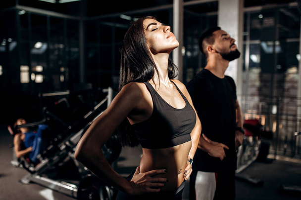 Slim dark-haired girl dressed in black sport clothes and fitness coach are doing warm up in the gym - 写真・画像