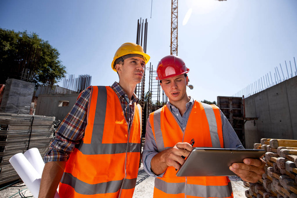 Architect  and structural engineer dressed in orange work vests and  helmets discuss a building project on the tablet on the open air building site with construction material - Photo, image