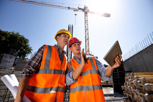 Architect  and structural engineer dressed in orange work vests and  helmets discuss a building project on the tablet on the open air building site with construction material - Фото, изображение